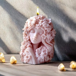 May include: A pink candle shaped like a woman's face with closed eyes and floral hair. The candle is lit, with a yellow flame at the top. The candle is on a wooden surface.