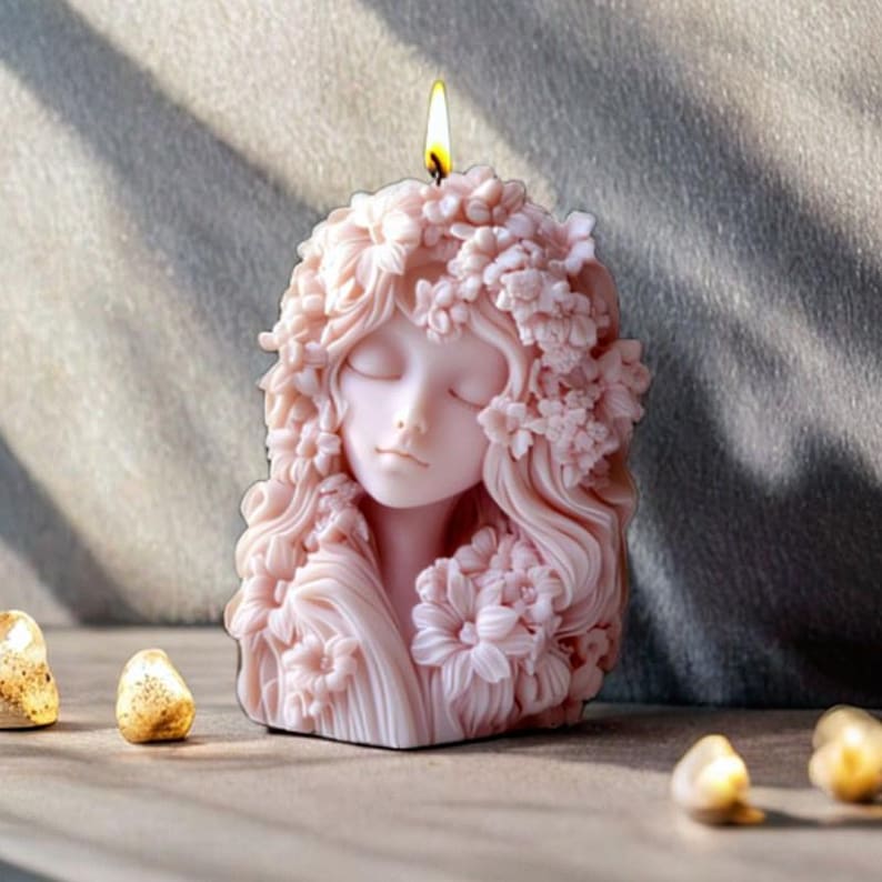 May include: A pink candle shaped like a woman's face with closed eyes and floral hair. The candle is lit, with a yellow flame at the top. The candle is on a wooden surface.