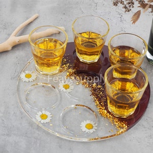 Peut inclure: Un plateau rond en résine marron avec six indentations circulaires, chacune contenant une marguerite blanche. Quatre verres à shot transparents remplis d'un liquide doré sont placés dans les indentations. Le plateau est décoré d'accents en feuilles d'or.