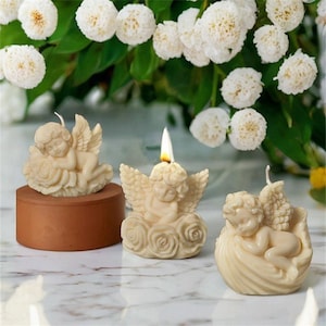 Puede incluir: Tres velas blancas con forma de ángel, cada una con una pose diferente. Un ángel está durmiendo sobre una rosa, otro está sentado sobre una nube de rosas y el tercero está durmiendo sobre una concha. Las velas son de cera y tienen un aspecto realista.