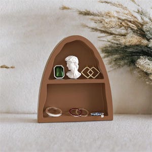 May include: A brown, arched shelf displaying jewellery and decorative items. The shelf holds a green gemstone, a white bust, and gold geometric earrings. Below are rings with clear, red, and blue accents. The shelf is a stylish storage solution.