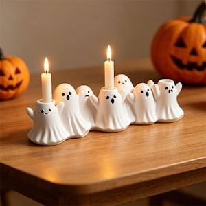 May include: A white ceramic candle holder shaped like a connected row of five ghosts with painted black eyes and mouths. Two thin, cream-colored taper candles are inserted into the holders, each emitting a warm, glowing flame from the lit wicks.