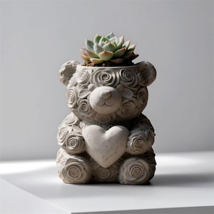May include: A gray concrete teddy bear planter with a heart on its chest, filled with a succulent plant. The bear is decorated with rose-shaped carvings. The succulent has green and pink leaves.