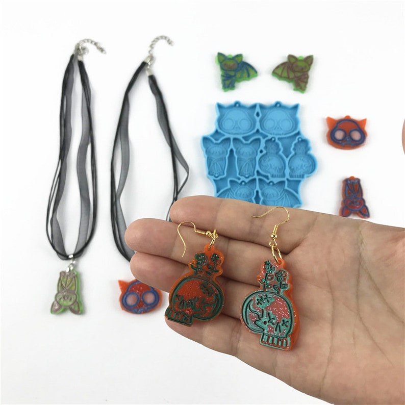 Puede incluir: Un conjunto de suministros para hacer joyas con tem&aacute;tica de Halloween, que incluye un molde de silicona azul con formas de calavera y murci&eacute;lago, dijes de resina naranja y verde, y un collar de cinta negra con un dije de murci&eacute;lago verde.