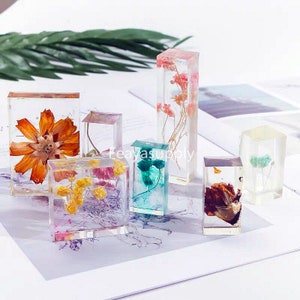 Pode incluir: Uma coleção de blocos de resina transparente, cada um com flores secas em várias cores. As flores incluem laranja, amarelo, rosa e azul-petróleo, criando uma exibição decorativa única. Os blocos estão dispostos sobre uma superfície branca.