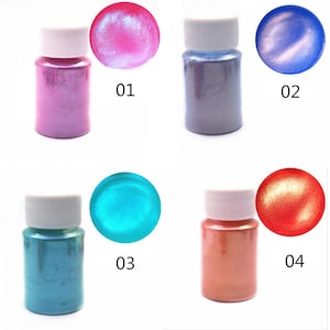 K&ouml;nnte beinhalten: Vier kleine Flaschen mit irisierendem Pigmentpulver in Rosa, Lila, Blau und Orange. Die Flaschen sind mit 01, 02, 03 und 04 beschriftet.