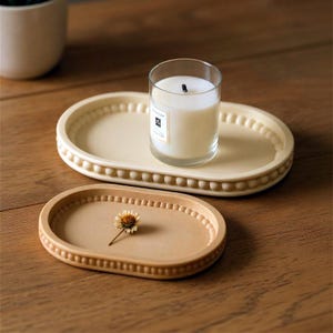 May include: Two oval decorative trays in cream and tan. The cream tray holds a clear glass candle holder with a lit candle. The tan tray has a small dried flower. Both trays have a beaded border.