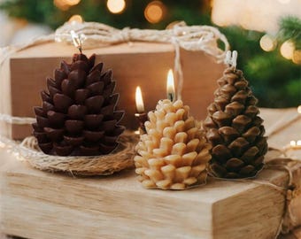 Puede incluir: Tres velas con forma de piña, en tonos marrones y beige, sobre una superficie de madera. Una vela está encendida, emitiendo un brillo cálido. Son velas decorativas, adecuadas para la decoración del hogar.