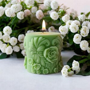 May include: A lit, green candle with floral relief carvings. The candle features detailed rose and leaf designs. The candle is surrounded by white flowers, creating a serene and decorative display. The candle's flame is visible.
