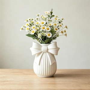 May include: A white ceramic vase with a bow design, filled with a bouquet of daisies. The vase has vertical ribbed detailing and sits on a wooden surface. The daisies have white petals and yellow centers.