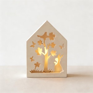 May include: A cream-coloured, house-shaped decorative light with cut-out designs of a tree, butterflies, a rabbit, and flowers. The interior glows with a warm light. Ideal for a nursery or as a decorative piece. The light is made of ceramic.