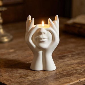 May include: A white ceramic candle holder shaped like a face with hands. The candle holder has a face with closed eyes and lips, with hands on either side of the face. A lit candle sits inside the holder, casting a warm glow.