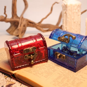 May include: Two small, clear plastic treasure chests with glitter. One chest is red and the other is blue. The blue chest is open and contains blue dice.