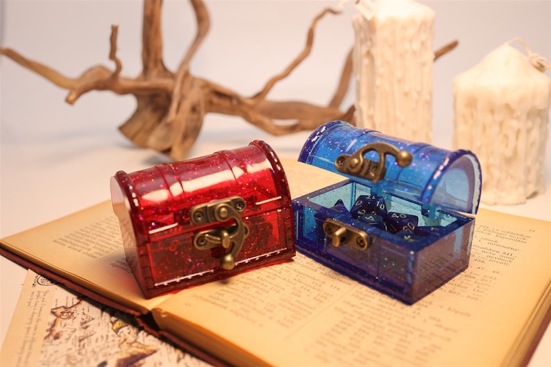 May include: Two small, clear plastic treasure chests with glitter. One chest is red and the other is blue. The blue chest is open and contains blue dice.