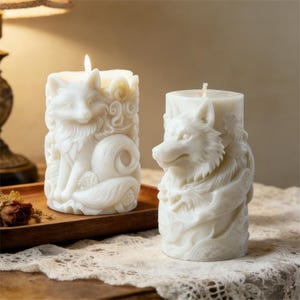 Puede incluir: Dos velas de pilar blancas sin perfume con tallas detalladas de animales. Una vela presenta un zorro, la otra un lobo. Ambas velas están encendidas, emitiendo un brillo cálido. Las velas están sobre una bandeja de madera y un mantel de encaje.