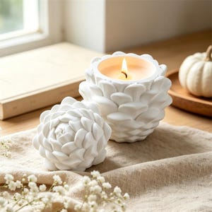 Peut inclure: Deux bougeoirs décoratifs blancs en forme de fleur. L'un est allumé avec une flamme chaude. L'autre est un couvercle. Les bougeoirs sont sur un tissu beige, avec un livre et une petite citrouille en arrière-plan.