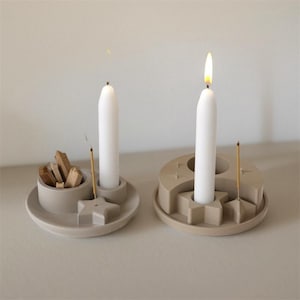 Peut inclure: Deux bougeoirs beiges avec des bougies blanches, dont une allumée. Les supports ont des formes d'étoiles et de croissants, avec un petit bol pour les bâtonnets d'encens et les copeaux de bois. Les bougies brûlent avec une flamme vive.