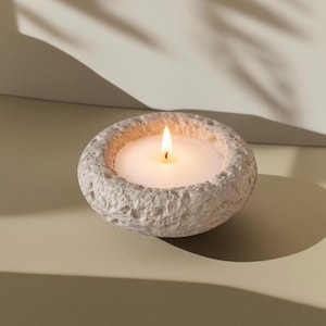 May include: A lit white candle in a textured, off-white stone bowl. The candle's flame is bright against the soft, neutral background. The bowl has a rough, organic texture, adding a natural element to the scene. The candle is a simple, elegant home decor item.