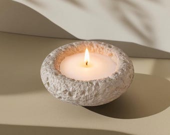 Puede incluir: Una vela blanca encendida en un cuenco de piedra texturizada de color blanquecino. La llama de la vela es brillante sobre el fondo suave y neutro. El cuenco tiene una textura rugosa y orgánica, añadiendo un elemento natural. Un artículo de decoración del hogar sencillo y elegante.