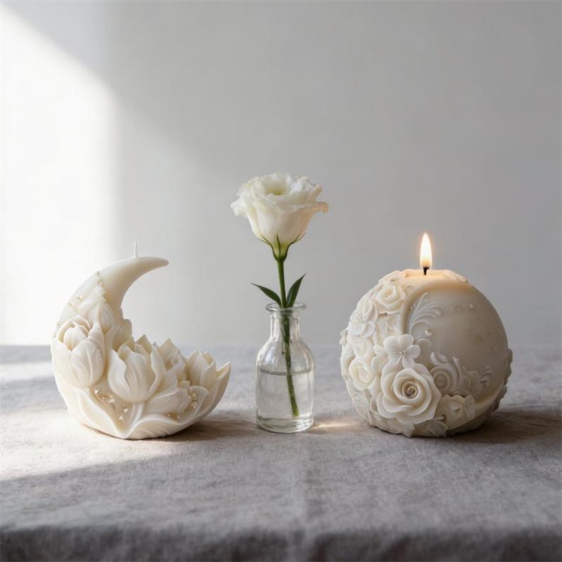 May include: A still-life composition featuring three decorative elements. A crescent moon-shaped candle with floral detailing, a white rose in a small glass vase, and a round candle with a floral design and a lit wick. The items are arranged on a gray surface.