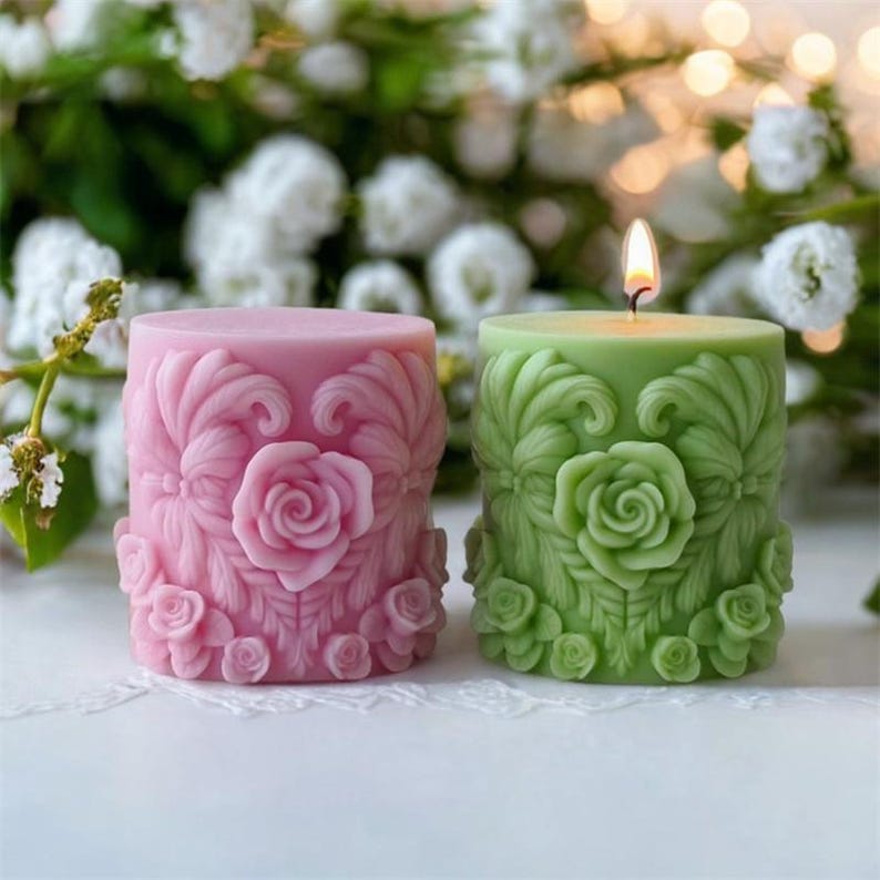 May include: Two decorative pillar candles. One is pink, the other is green, both with a floral rose design. The pink candle is unlit, while the green candle has a lit wick. The candles are surrounded by white flowers.