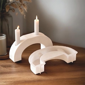 May include: Two white, arch-shaped candle holders with lit candles. One holder has two candles, the other is empty. The holders are on a wooden surface, creating a minimalist, modern aesthetic.