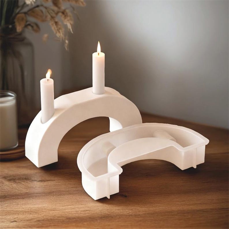 May include: Two white, arch-shaped candle holders with lit candles. One holder has two candles, the other is empty. The holders are on a wooden surface, creating a minimalist, modern aesthetic.