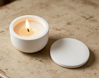 May include: A lit white candle in a round, white ceramic container with a matching lid. The candle flame is bright against the creamy wax. The lid is next to the candle on a distressed, light-colored wooden surface.