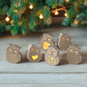 Puede incluir: Seis luces decorativas con forma de búho marrón. Cada búho tiene ojos grandes y redondos y un diseño recortado en el pecho, con un cálido brillo amarillo que emana del interior. Las luces están dispuestas sobre una superficie de madera.