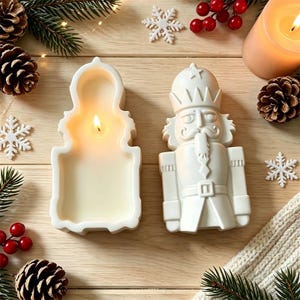 May include: A white nutcracker-shaped candle and a matching white nutcracker-shaped candle mold. The candle is lit, showing a flame inside the hollow form. The set is arranged with pine cones, red berries, and white snowflake ornaments.