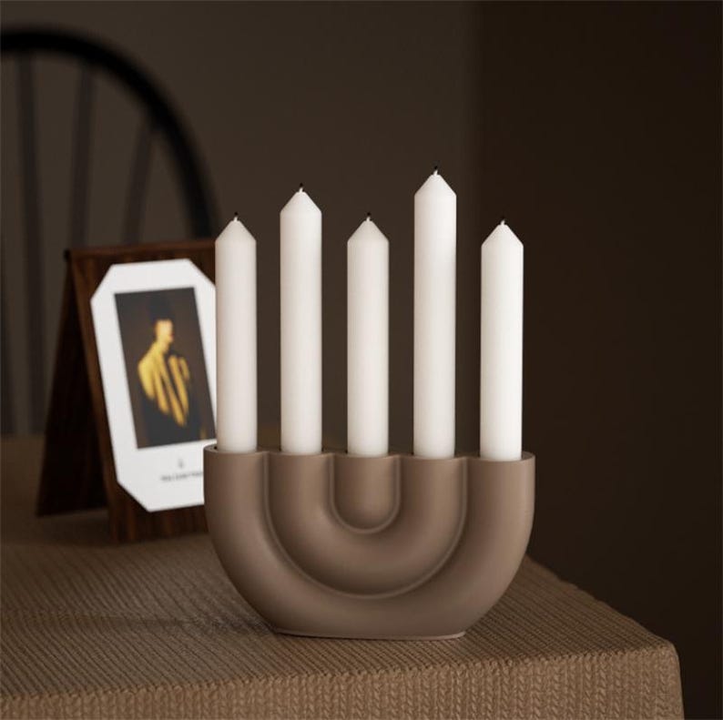 May include: A brown ceramic candle holder with five white candles. The holder has a curved design and sits on a textured surface. A framed picture is in the background.