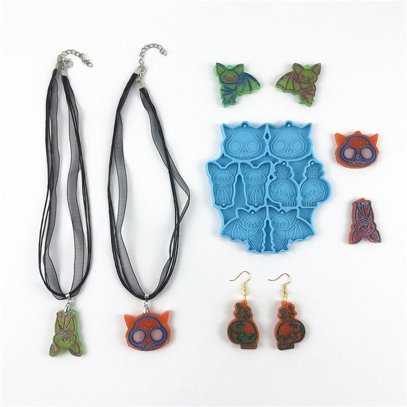 Puede incluir: Un conjunto de suministros para hacer joyas con tem&aacute;tica de Halloween, que incluye dos collares de cinta negra, un molde de silicona azul con formas de murci&eacute;lago y calavera, y pendientes y colgantes de resina naranja y verde.