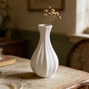 May include: A white ceramic vase with a fluted design, holding dried flowers. The vase has a narrow neck and a bulbous base. It sits on a vintage table. The overall aesthetic is simple and elegant.