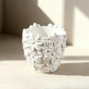 May include: White ceramic flower pot with a detailed floral design. The pot is cylindrical with a rim of flower petals. The exterior is covered in raised, three-dimensional flowers of varying sizes. The pot is suitable for indoor or outdoor use.