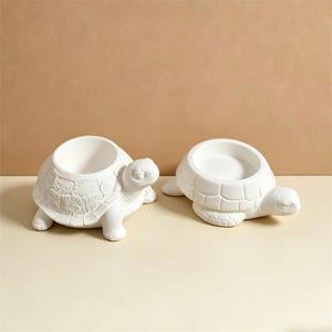 May include: Two white ceramic turtle-shaped planters. Each planter has a round opening for plants or other items. The turtles have detailed shell patterns and are on a light-coloured surface.