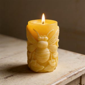 May include: A lit, yellow beeswax candle with a carved bee design. The candle is cylindrical and has a detailed relief of a bee with wings and leaves. The flame is bright against the warm yellow wax, creating a cozy ambiance.