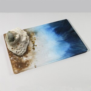 May include: A rectangular tray with a blue and white ocean-inspired design. The tray has a brown sand-like texture at the bottom and a white seashell on top.