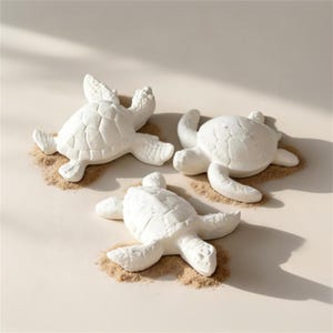 Op de afbeelding: Drie witte keramische schildpadfiguren die op kleine stukjes zand liggen. De schildpadden hebben gedetailleerde schaalpatronen en uitgestrekte vinnen, wat een decoratief item met een kust- of strandthema creëert.