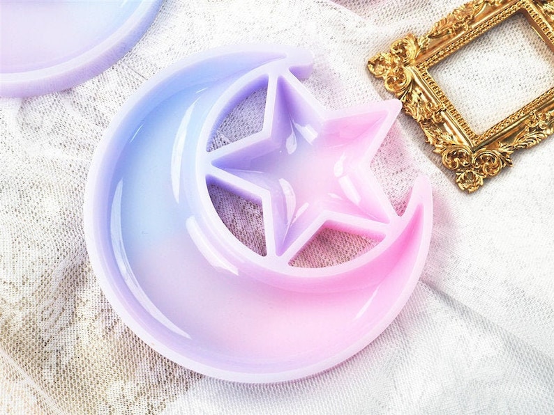 May include: A light purple and pink crescent moon shaped resin mold with a five-pointed star cutout in the center. The mold is perfect for creating unique resin crafts.