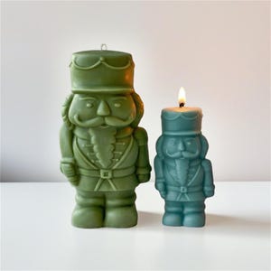Könnte beinhalten: Zwei dekorative Kerzen in Form von Nussknacker-Soldaten. Eine ist ein größeres Olivgrün, die andere ein kleineres Türkisblau mit einer brennenden Flamme. Beide Kerzen haben detaillierte Gesichtszüge und Militäruniformen.
