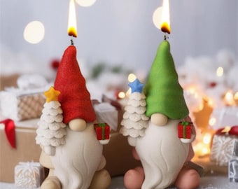 Puede incluir: Dos velas de gnomo de Navidad con un sombrero rojo y verde, sosteniendo un pequeño árbol de Navidad blanco con una estrella en la parte superior. Las velas están encendidas y tienen una barba blanca y una caja de regalo roja y verde.