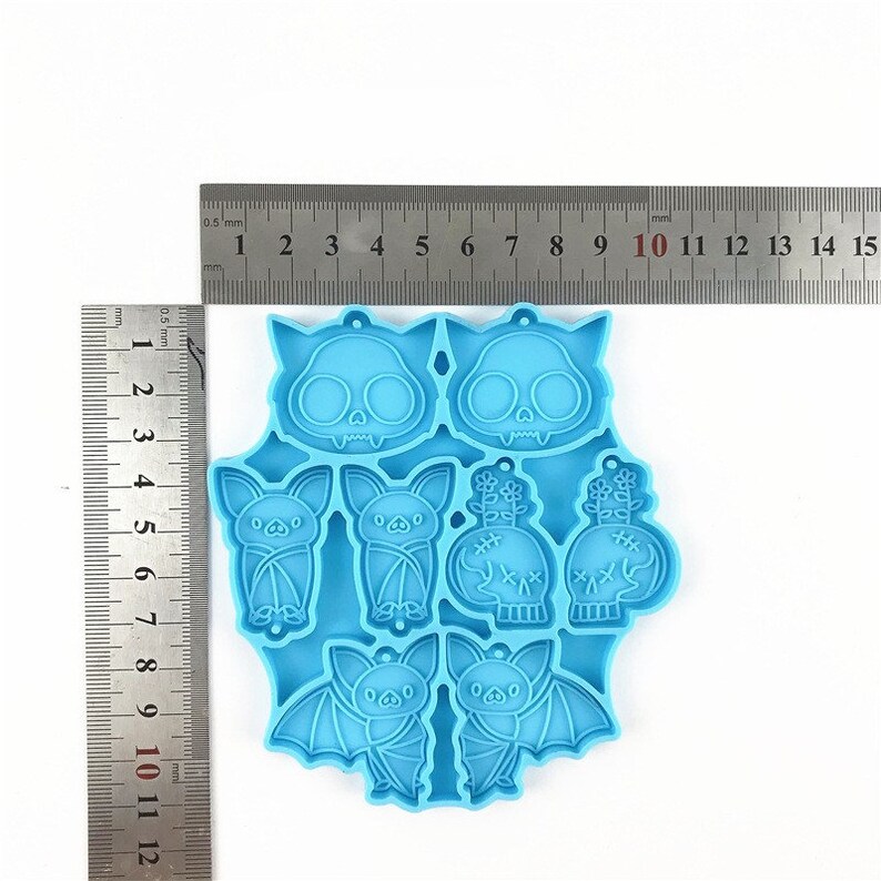 Puede incluir: Molde de silicona azul con ocho formas de tem&aacute;tica de Halloween: dos murci&eacute;lagos, dos calaveras con huesos cruzados, dos murci&eacute;lagos con las alas plegadas y dos calaveras con flores. El molde est&aacute; sobre una superficie blanca con una regla para la escala.