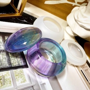 Puede incluir: Un juego de dos platos para anillos de resina iridiscente con tapa. Los platos son de color azul claro y morado con un acabado brillante. La tapa es de color azul claro con un acabado brillante.