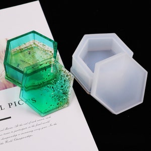 Puede incluir: Dos moldes hexagonales de silicona para hacer cajas de joyería de resina. Un molde está vacío y el otro está lleno de resina verde con purpurina dorada.