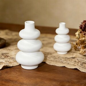 May include: Two white ceramic vases with a stacked, rounded design. The vases have a smooth, matte finish and a small opening at the top. They are placed on a wooden surface with a patterned runner.