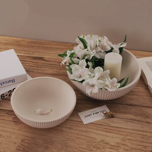 May include: Two white ceramic bowls with a ribbed texture. The larger bowl is filled with white flowers and a white candle. The smaller bowl is empty. The bowls are on a wooden surface with a card that says "Just for you".
