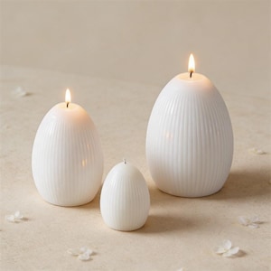 Può includere: Tre candele bianche a forma di uovo con dettagli a coste verticali. Due candele sono accese, con una piccola fiamma in cima. Le candele variano di dimensioni, creando un'esposizione decorativa. Lo sfondo è di colore neutro.