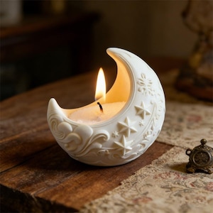 Peut inclure: Un bougeoir blanc en forme de croissant de lune avec une bougie allumée. Le bougeoir présente des étoiles, des nuages et des motifs floraux en relief. La bougie brûle avec une flamme vive, dégageant une lueur chaleureuse. Le bougeoir est sur une surface en bois.