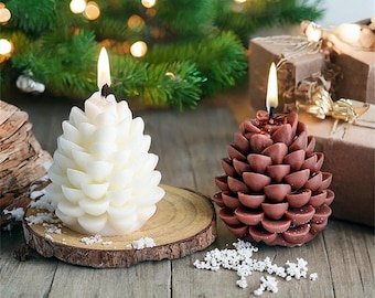 May include: Two pine cone shaped candles, one white and one brown, on a wooden slice. The candles are on a wooden table with white snowflake confetti.