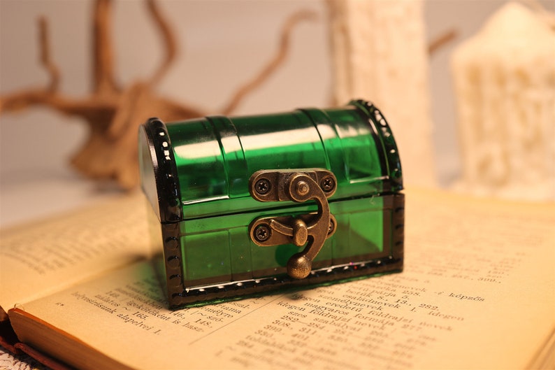 May include: A small, green, plastic treasure chest with a brass latch and hinges. The chest is sitting on top of an open book.